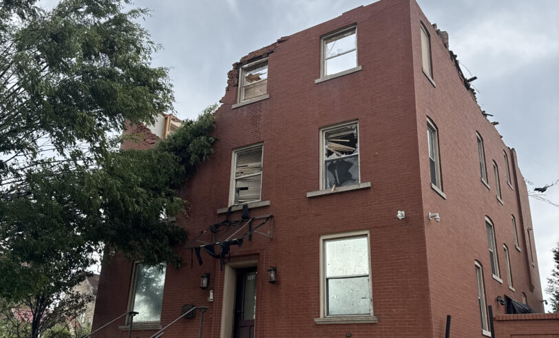 Damaged red brick office building with missing roof and third floor after a tornado, shattered windows, and debris around the entrance.